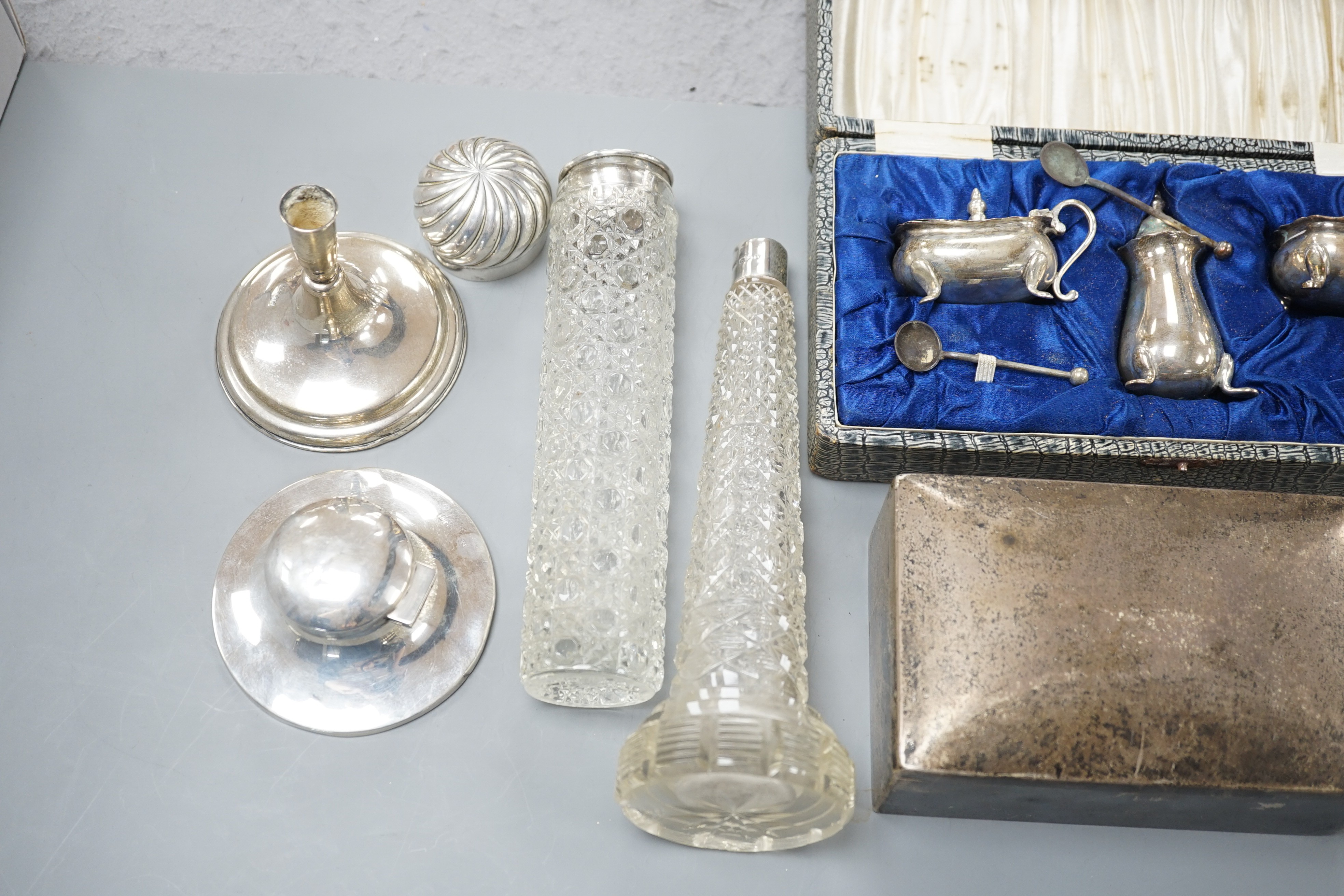 Assorted silver items including a cigarette box, a cased silver condiment set, two capstan inkwells, a pair of Edwardian dwarf candlesticks and four silver mounted glass toilet jars.